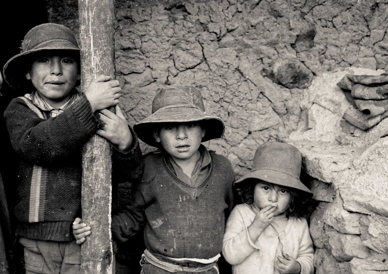 Trois enfants coiffés de chapeaux de feutre devant un mur de terre, en noir et blanc.