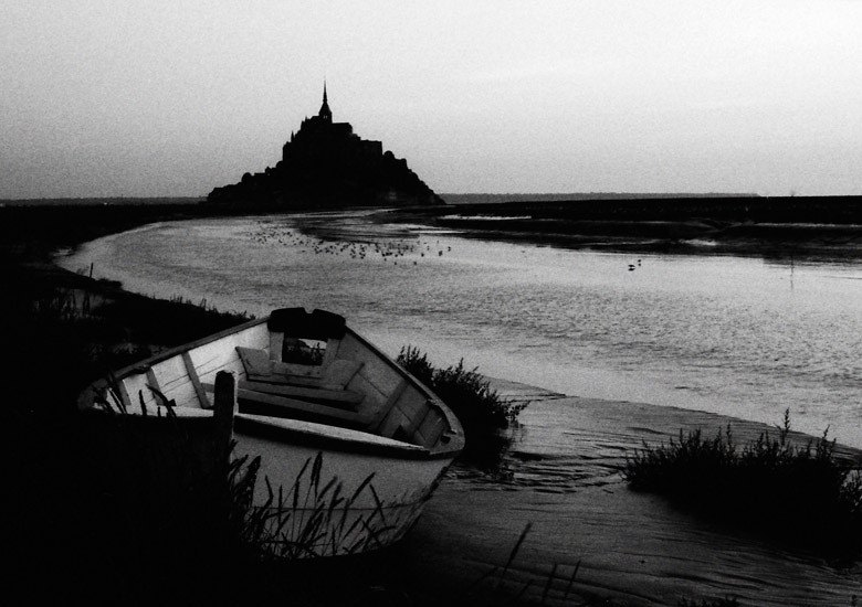 Le Mont-Saint-Michel au crépuscule, en noir et blanc, une barque échouée sur la grève au premier plan.